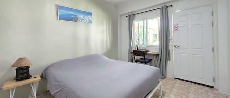 Ein Schlafzimmer mit Bett, Schreibtisch, Stuhl, Lampe, einem Fenster mit Vorhängen und einer Tür.