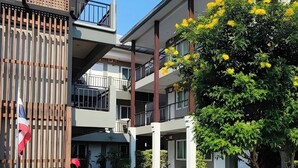 Un edificio de varios pisos con balcones, una bandera y un árbol con flores amarillas.