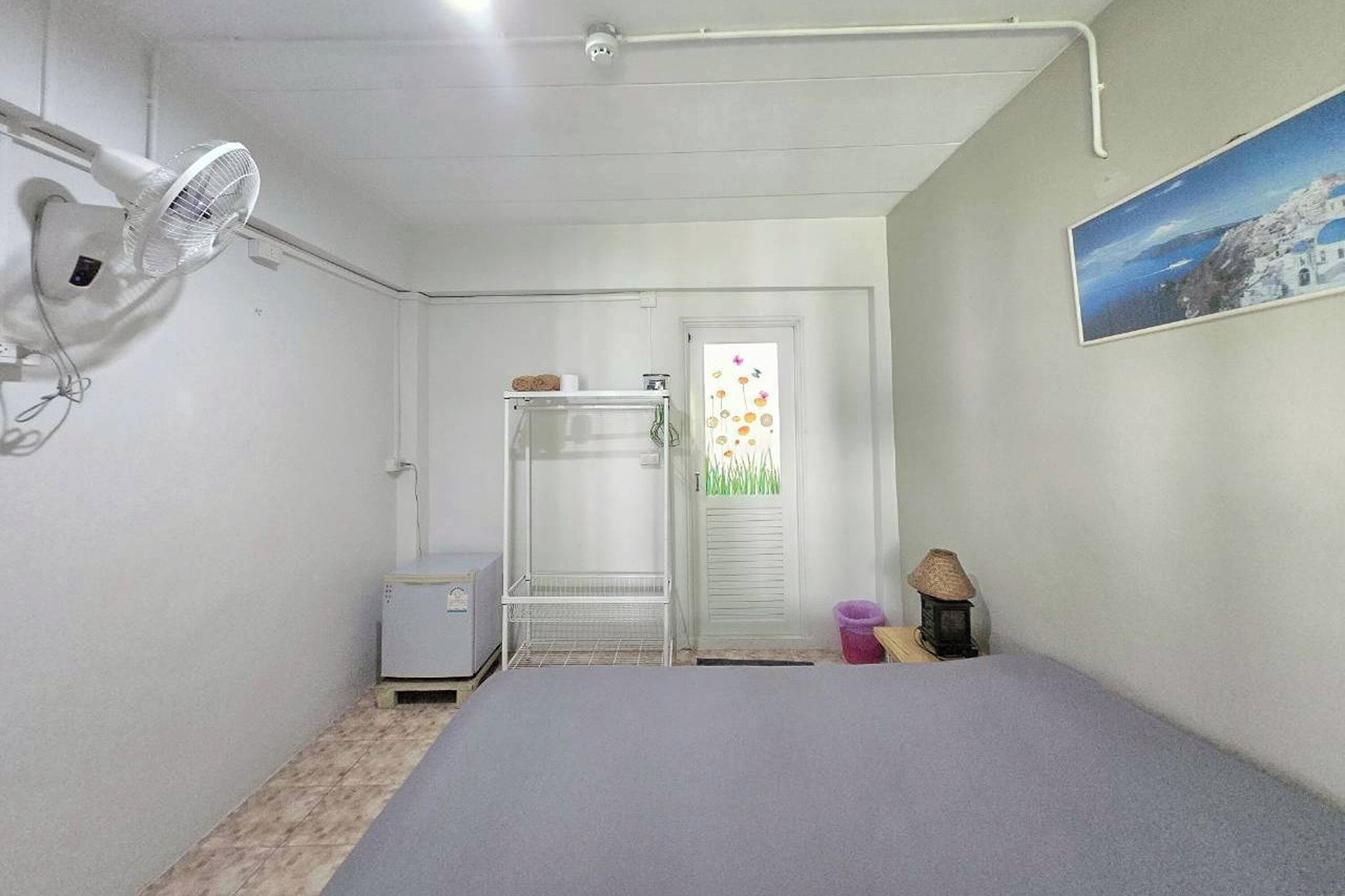 Une petite pièce simple, avec un lit, un ventilateur, un petit radiateur et une porte donnant sur une autre pièce.