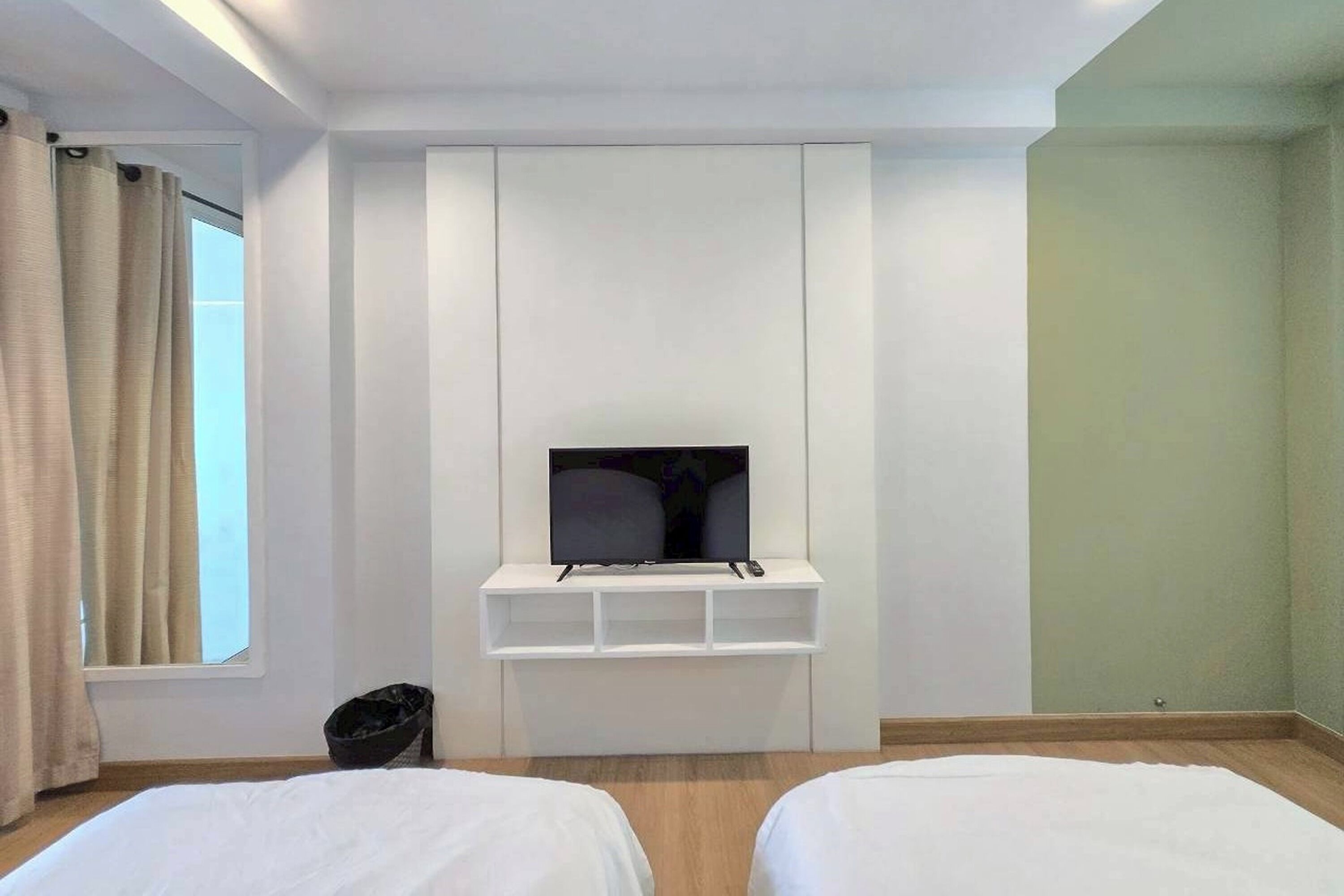 A hotel room with a TV mounted on a white wall, two beds with white bedding, a small trash bin, and a window with beige curtains.