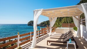 A seaside terrace with a wooden deck, white curtains, a lounge chair, and a pool. - Villa Renata & Villa Filippos with private Plunge Pool by Konnect (Corfu)