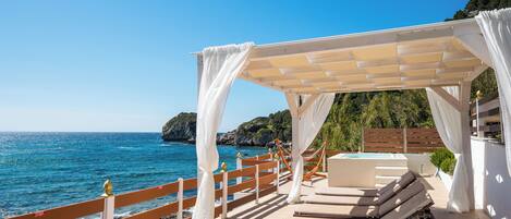 A seaside terrace with a wooden deck, white curtains, a lounge chair, and a pool.