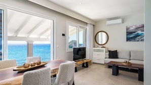 A modern living room with a large window overlooking the sea, a dining area with a wooden table and chairs, and a sofa with a side table. - Villa Renata & Villa Filippos with private Plunge Pool by Konnect (Corfu)