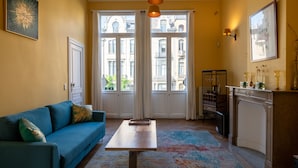 Una habitación con sofá azul, mesa de madera, alfombra colorida, zona de cocina y ventanas grandes con vistas a edificios.