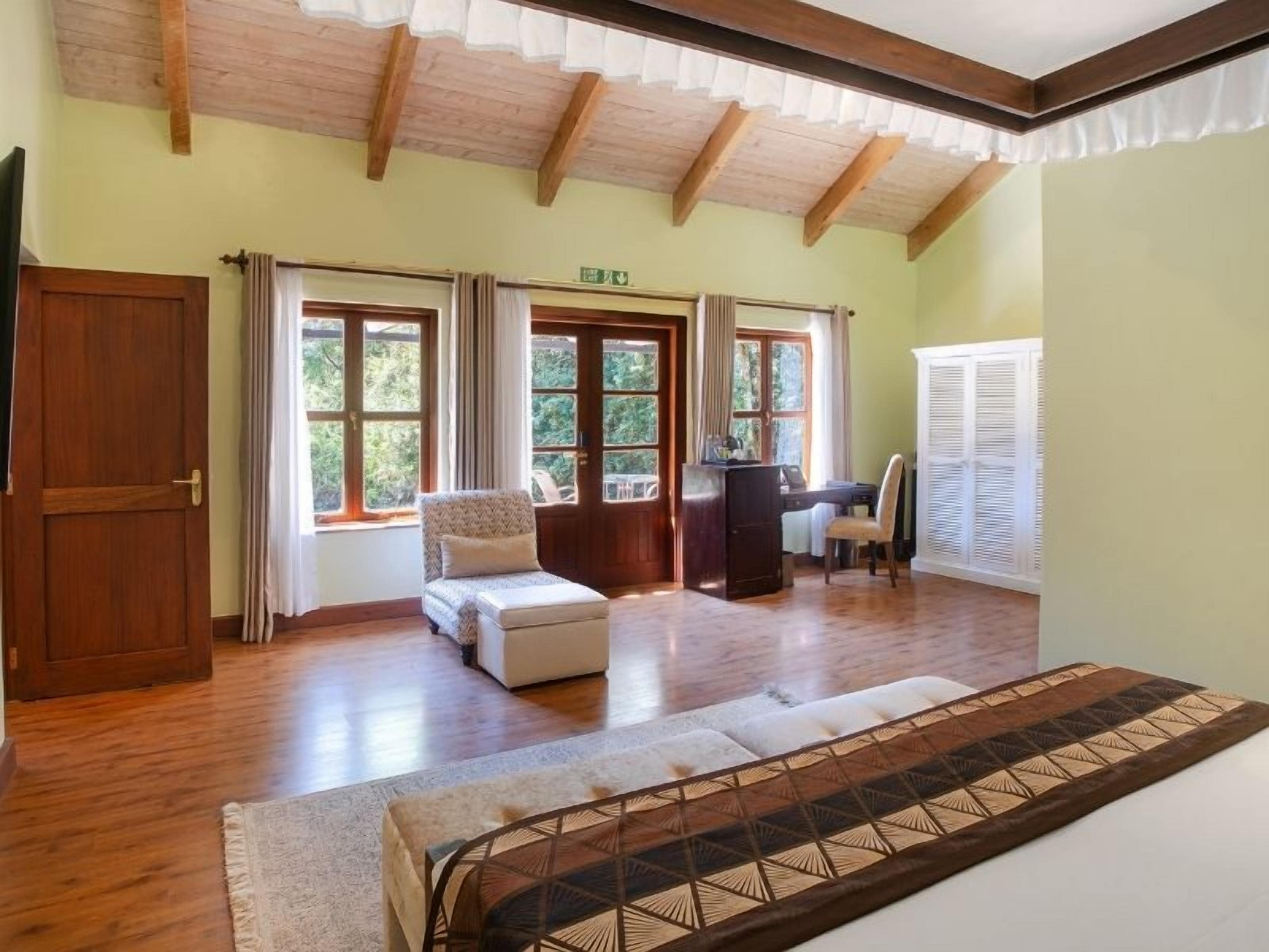 A room with a bed, a desk, and a chair. There are windows with curtains, a wooden door, and a ceiling with exposed beams.