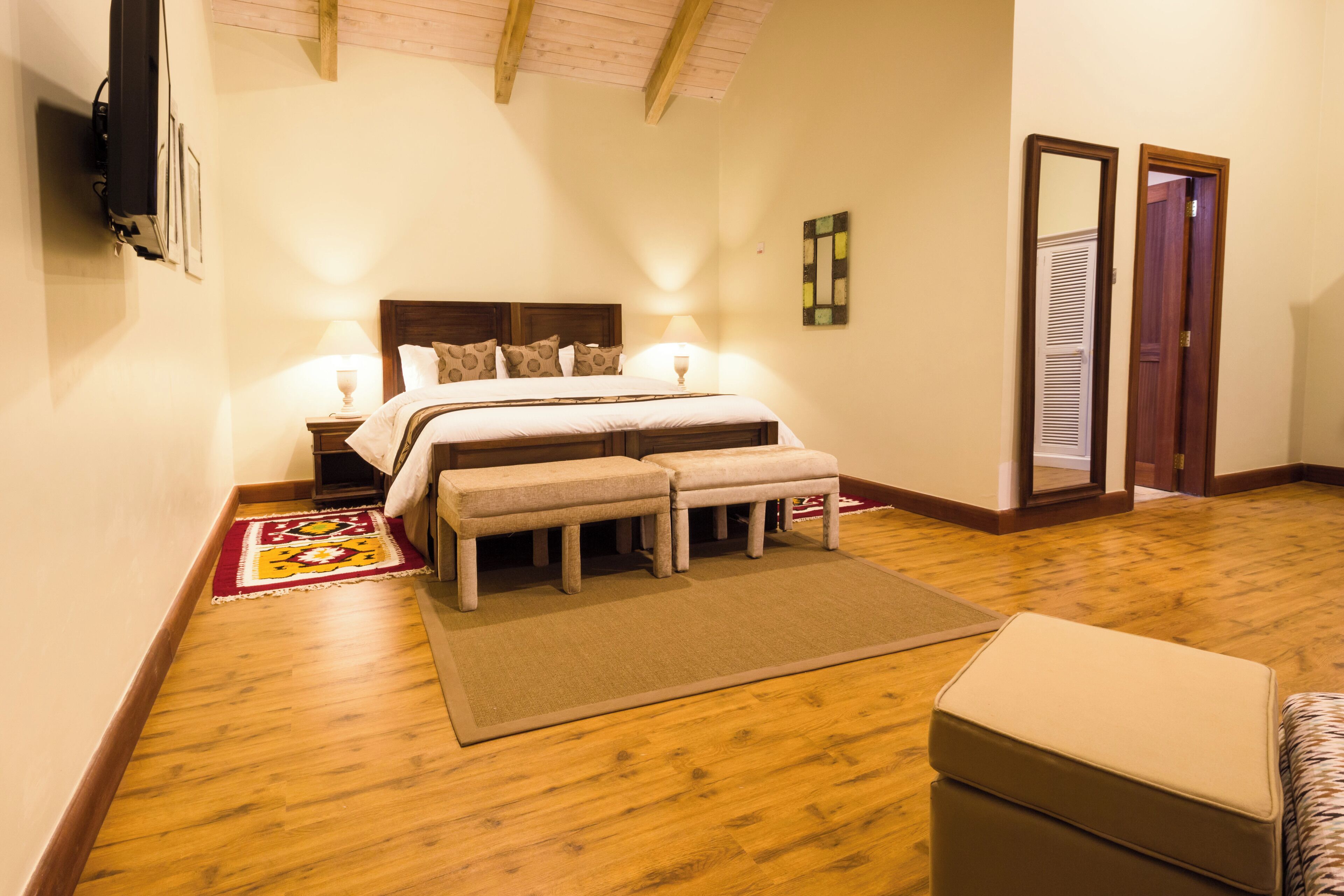 A bedroom with a wooden floor, a bed with two pillows, a bench, and a bedside table with a lamp.