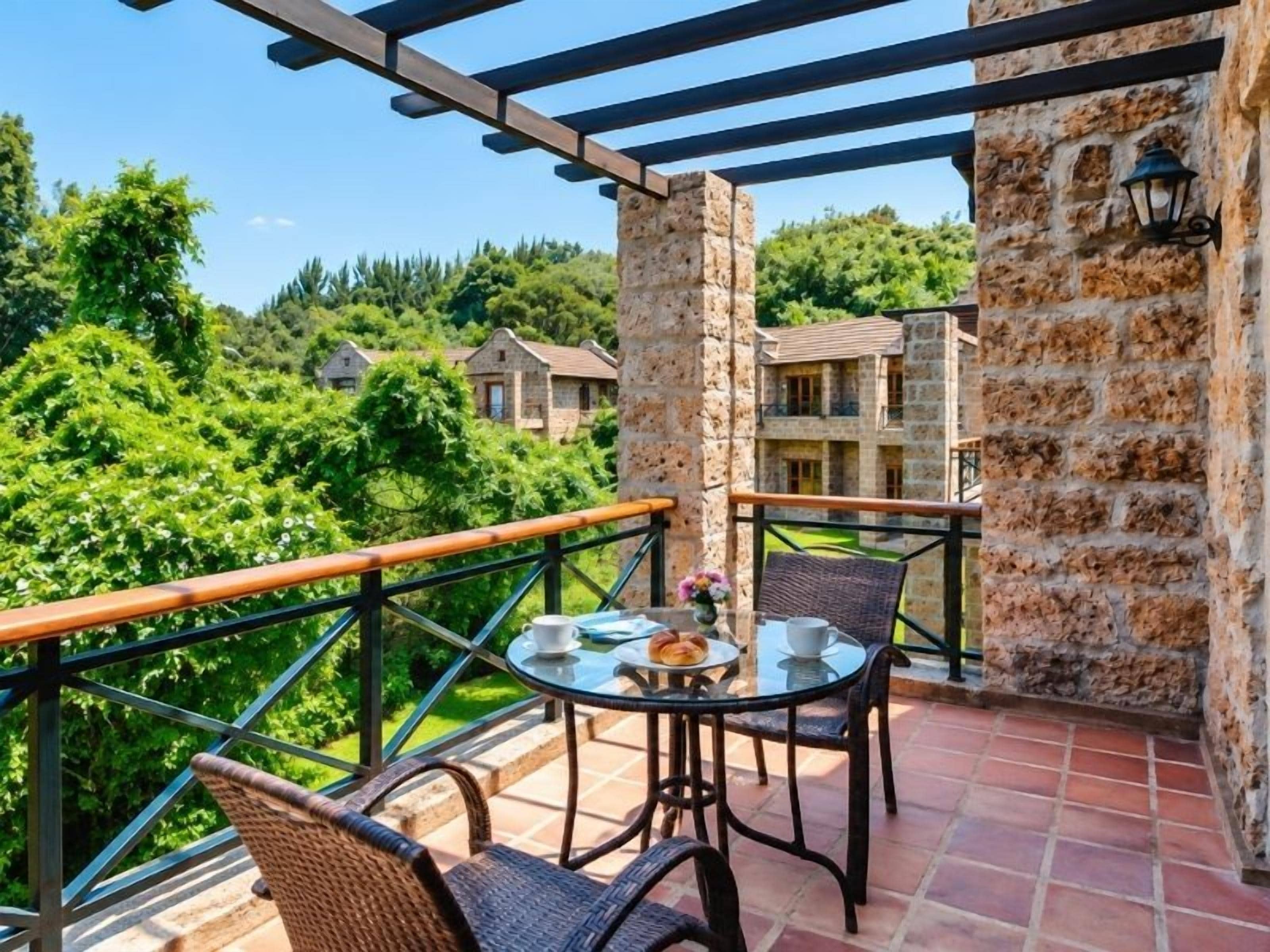 A balcony with a table and chairs, a stone wall, and a view of a building and greenery.