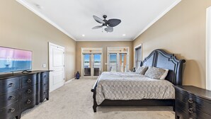 Une chambre à coucher comprenant un lit, une commode, un téléviseur, un ventilateur de plafond et deux portes donnant sur un balcon.