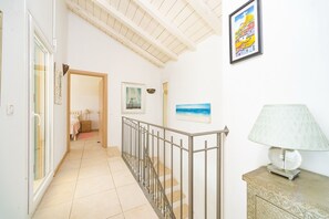 A hallway with a staircase, a bed in the background, and a lamp on a cabinet.