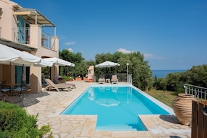 A swimming pool with lounge chairs and umbrellas, surrounded by stone flooring and greenery, with a view of the sea.