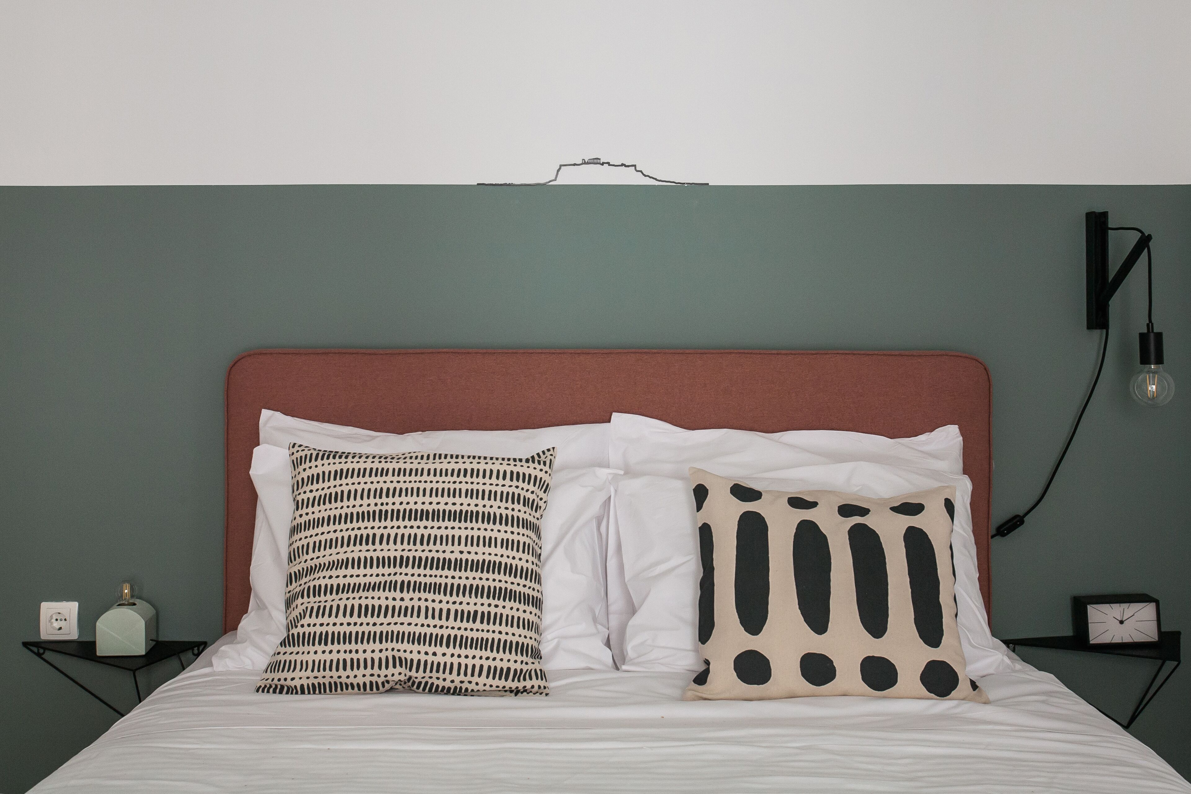 Una cama con dos almohadas, una mesita de noche con un reloj y una lámpara fijada a la pared.