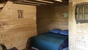 A cozy wooden cabin room with a bed, a chair, a window, and a wall-mounted lamp. - Charming tree house in Normandy (Essay)
