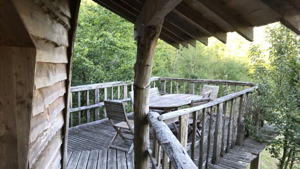 A wooden deck with a table and chairs, surrounded by a wooden railing, overlooking a forested area. - Charming tree house in Normandy (Essay)