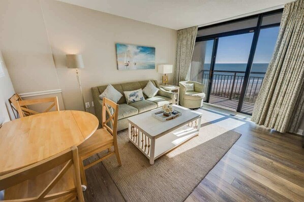 A modern living room with a sofa, coffee table, and chairs. There is a painting on the wall and a view of the ocean through large windows. - WOW Newly Updated Ocean Front Condo with New Pools (North Myrtle Beach)