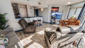 A modern living room with a sofa, a recliner, a dining area with a table and chairs, a kitchen area with a bar, and a large window with a view. - 1BR Oceanview at Carolinian, Pools, Amazing Views! (Myrtle Beach)