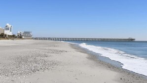 A beach with a long pier extending into the water, a building with a white facade, and a clear blue sky. - 1BR Oceanview at Carolinian, Pools, Amazing Views! (Myrtle Beach)
