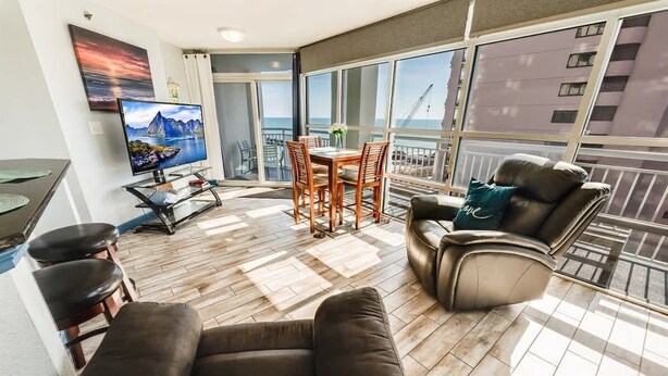 A modern living room with a flat-screen TV, a dining table, and a sofa. - 1BR Oceanview at Carolinian, Pools, Amazing Views! (Myrtle Beach)