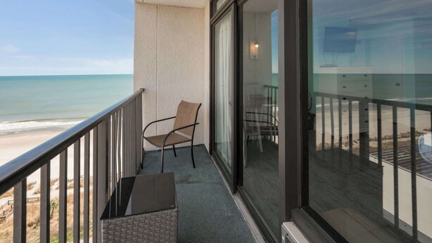 A balcony with a view of the beach, a chair, and a table.