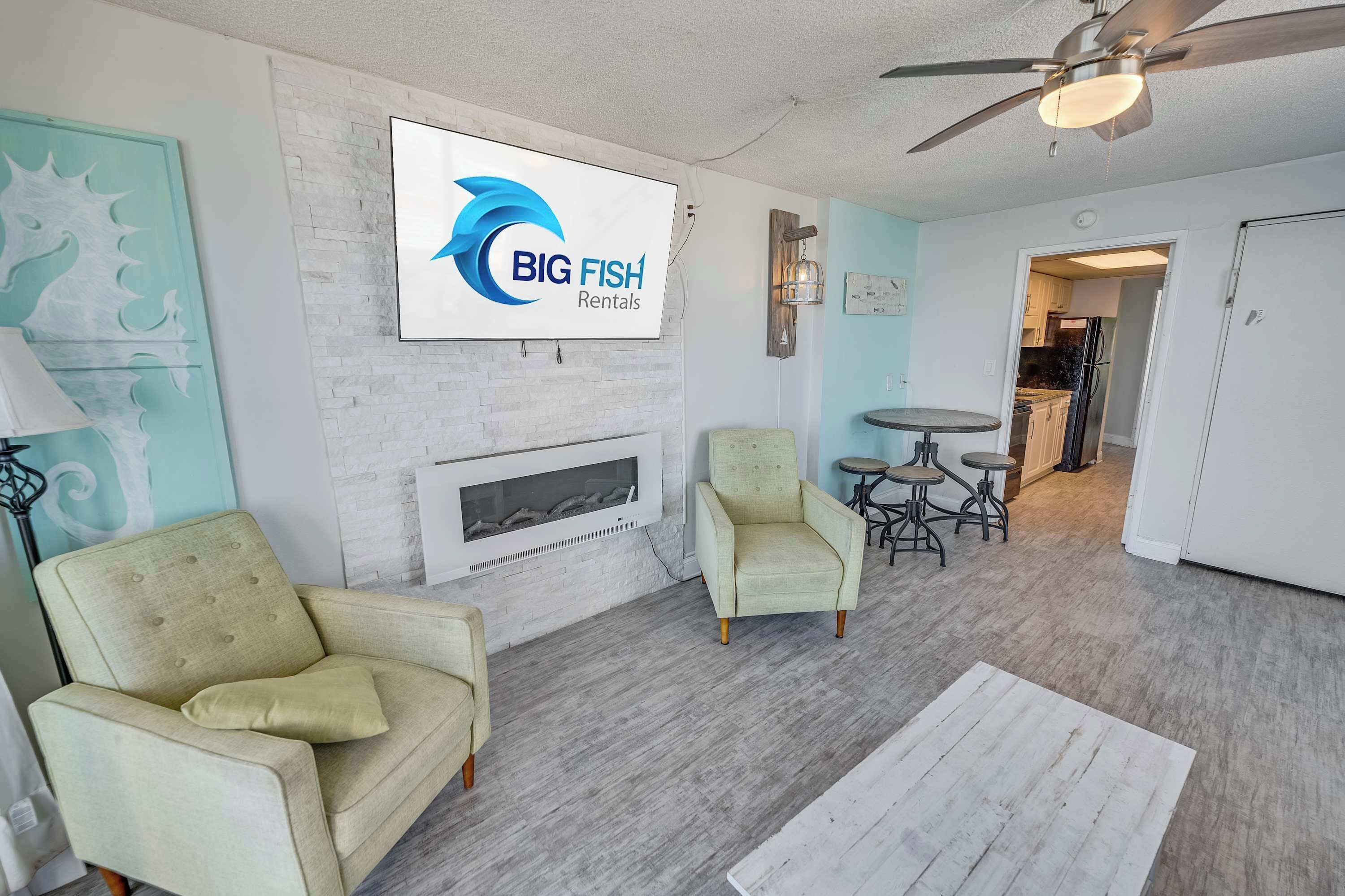 Un vaste espace de vie doté d’une cheminée, de deux fauteuils, d’une table à manger et d’un ventilateur de plafond. Une enseigne indiquant « BIG FISH Rentals » est visible sur le mur.