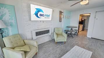 Un vaste espace de vie doté d’une cheminée, de deux fauteuils, d’une table à manger et d’un ventilateur de plafond. Une enseigne indiquant « BIG FISH Rentals » est visible sur le mur.