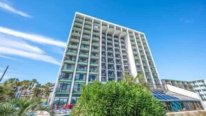 A high-rise hotel with multiple balconies, surrounded by greenery and a clear blue sky. - Pets welcome,direct oceanfront, poolside,1 bedroom (Myrtle Beach)