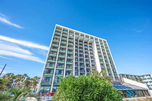 A high-rise hotel with multiple balconies, surrounded by greenery and a clear blue sky.