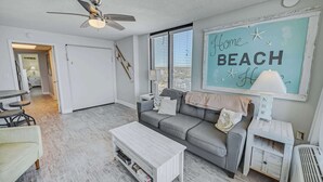 A living room with a grey sofa, a white coffee table, a ceiling fan, and a large window with blinds. - Pets welcome,direct oceanfront, poolside,1 bedroom (Myrtle Beach)