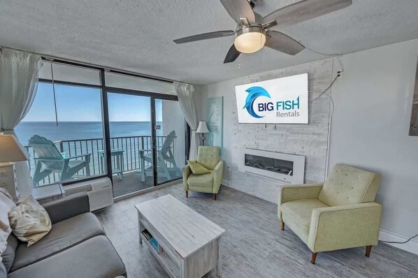 A spacious living room with a large sliding glass door leading to a balcony overlooking the sea. There is a ceiling fan, a fireplace, and two armchairs.