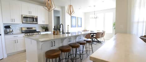 A modern kitchen with white cabinets, a large island with stools, a built-in oven, and pendant lighting.