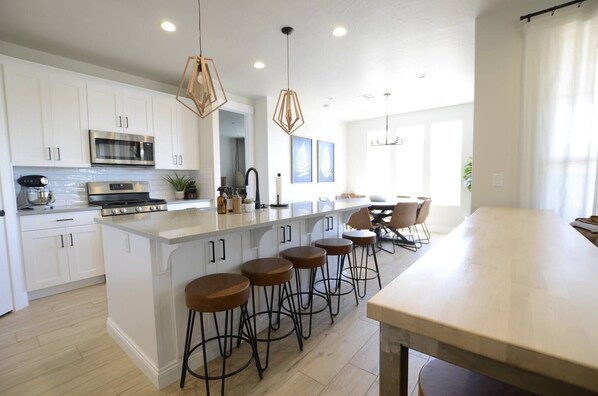 A modern kitchen with white cabinets, a large island with stools, a built-in oven, and pendant lighting.