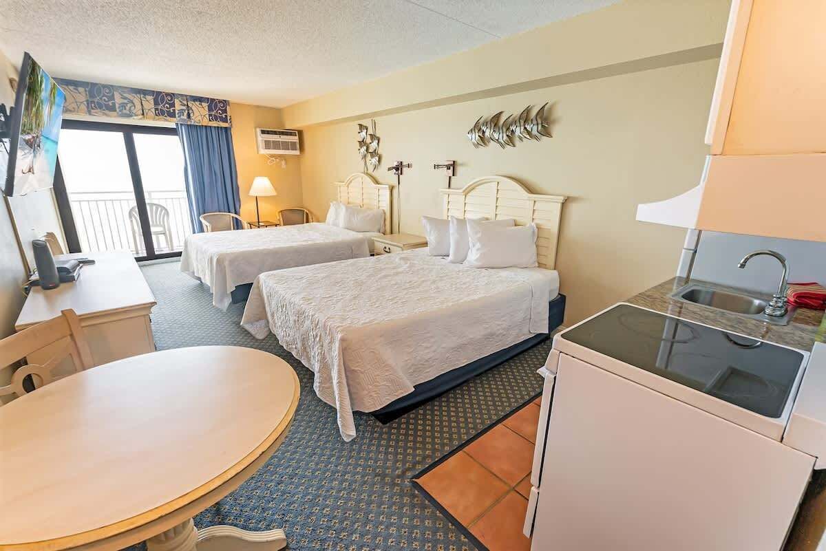 A hotel room with two beds, a desk, a chair, a small table, a sink, and a balcony with a view.