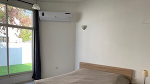 Une chambre à coucher comprenant un lit, une table de chevet, une lampe, un appareil de climatisation et une fenêtre avec des rideaux.