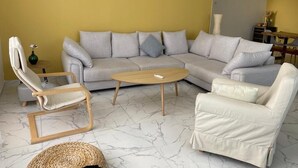 Un salon comprenant un canapé d’angle gris, une table basse en bois, un fauteuil blanc et un pouf en osier.