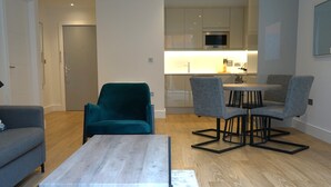 Een moderne woonkamer met een grijze bank, een blauwgroene fauteuil en een salontafel. In de achtergrond is een keuken met witte kasten en een magnetron zichtbaar.