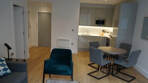 Een moderne woonkamer met een bank, een blauwgroene fauteuil en een eettafel met vier stoelen. In de achtergrond is een keuken met een magnetron en kasten zichtbaar.
