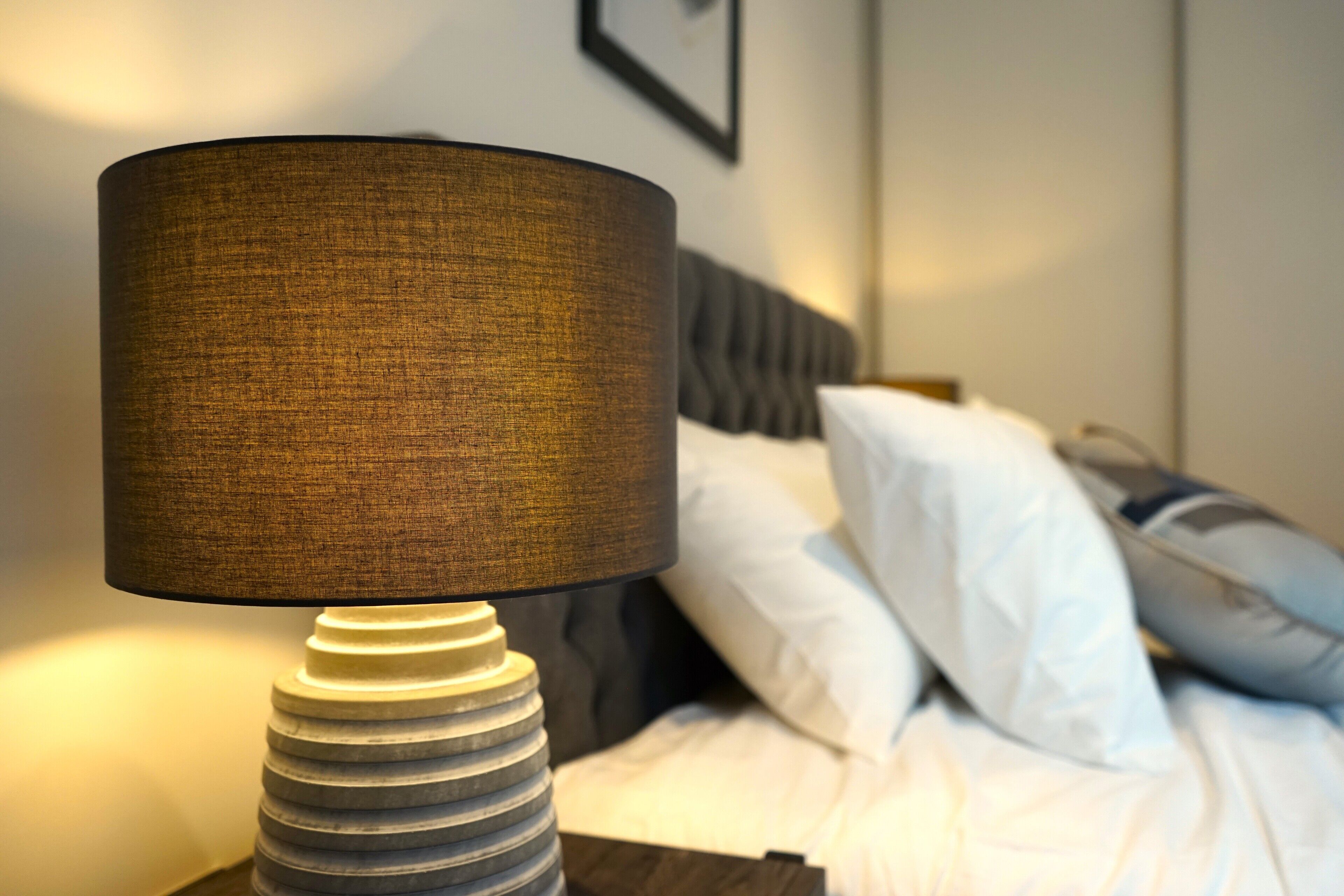 Ein Bett mit weißer Bettwäsche und einem grauen Kopfteil, eine Nachttischlampe mit einer strukturierten Lampe und ein gerahmtes Bild an der Wand.