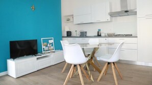 Uma cozinha moderna com armários brancos, uma sala de jantar com mesa redonda e cadeiras brancas, e uma TV sobre um móvel branco.