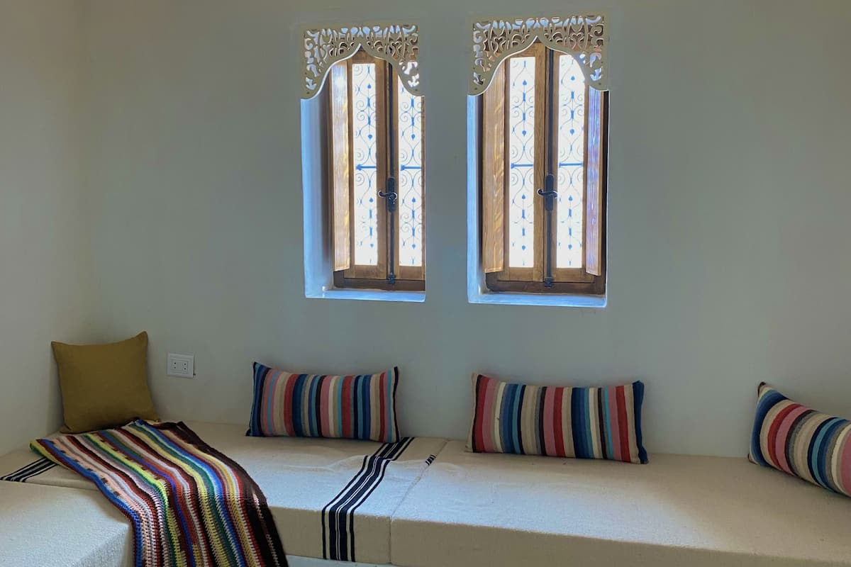 A room with a white wall, two windows with decorative grills, a white sofa with colorful striped cushions, and a multicolored striped rug.