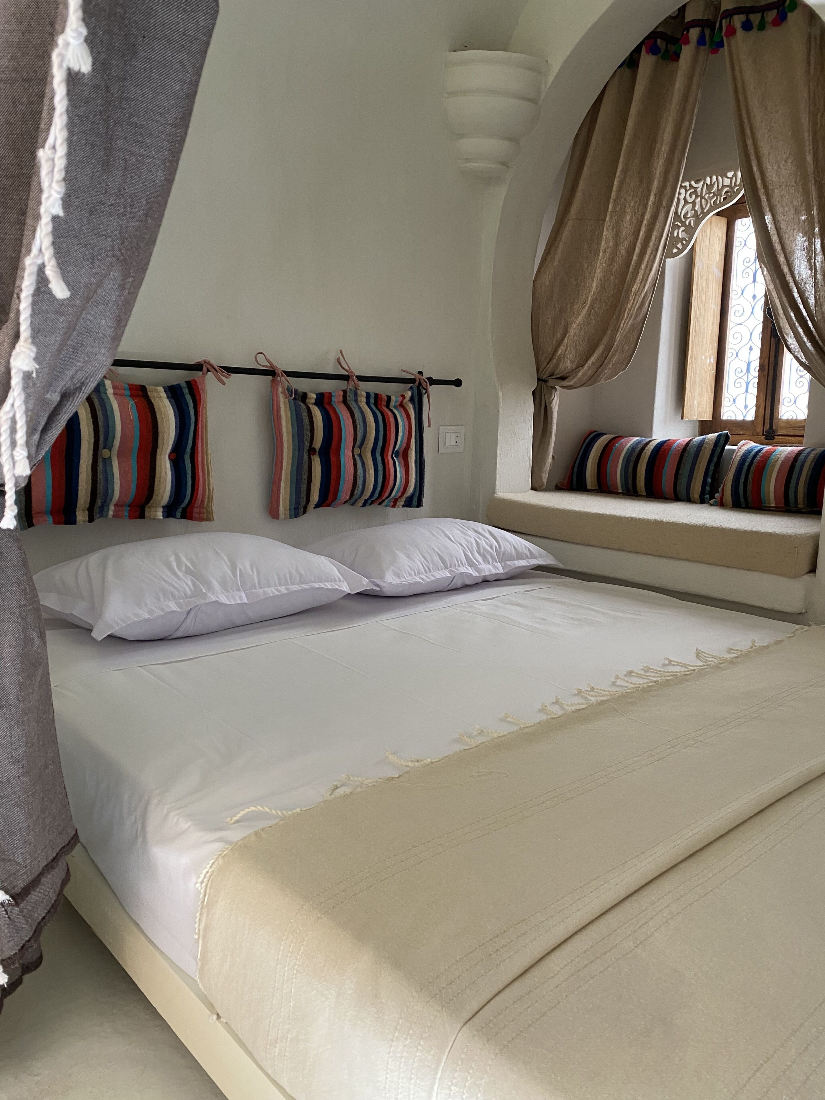 Een bed met witte lakens, een beige deken en gestreepte kussens. Er is een raam met gordijnen en uitzicht op de omgeving.