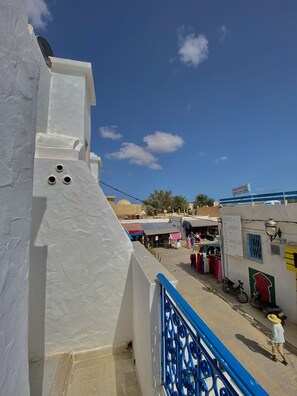 A white building with a blue railing, a clear blue sky, and a street with shops and people.