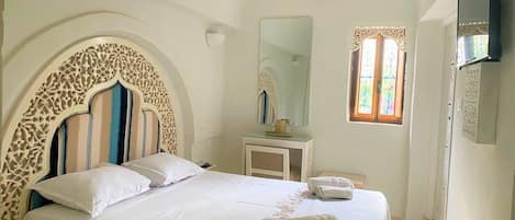 A bedroom with a large bed, a bedside table, a mirror, and a window with a stained glass design.