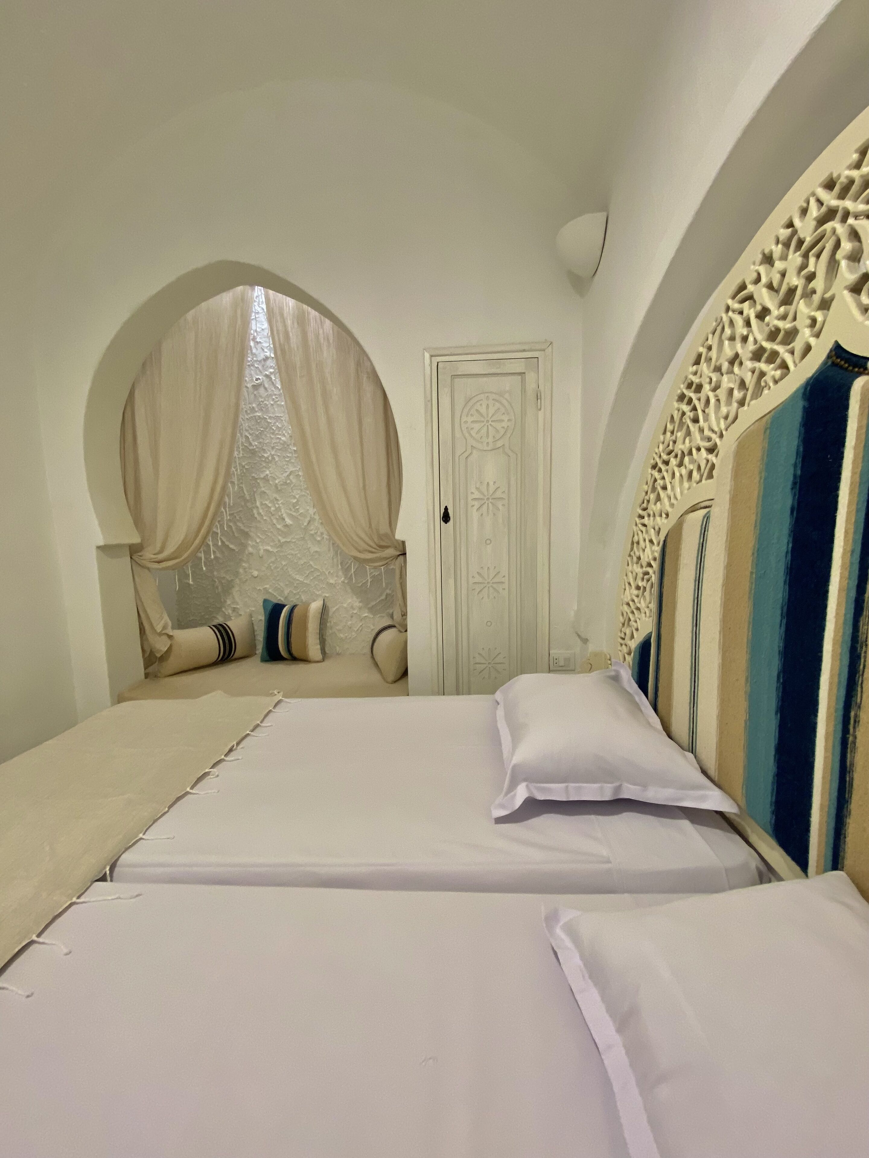 Ein Schlafzimmer mit zwei Betten, einem dekorativen Rundbogenfenster und einer weißen Tür mit aufwendigen Verzierungen.
