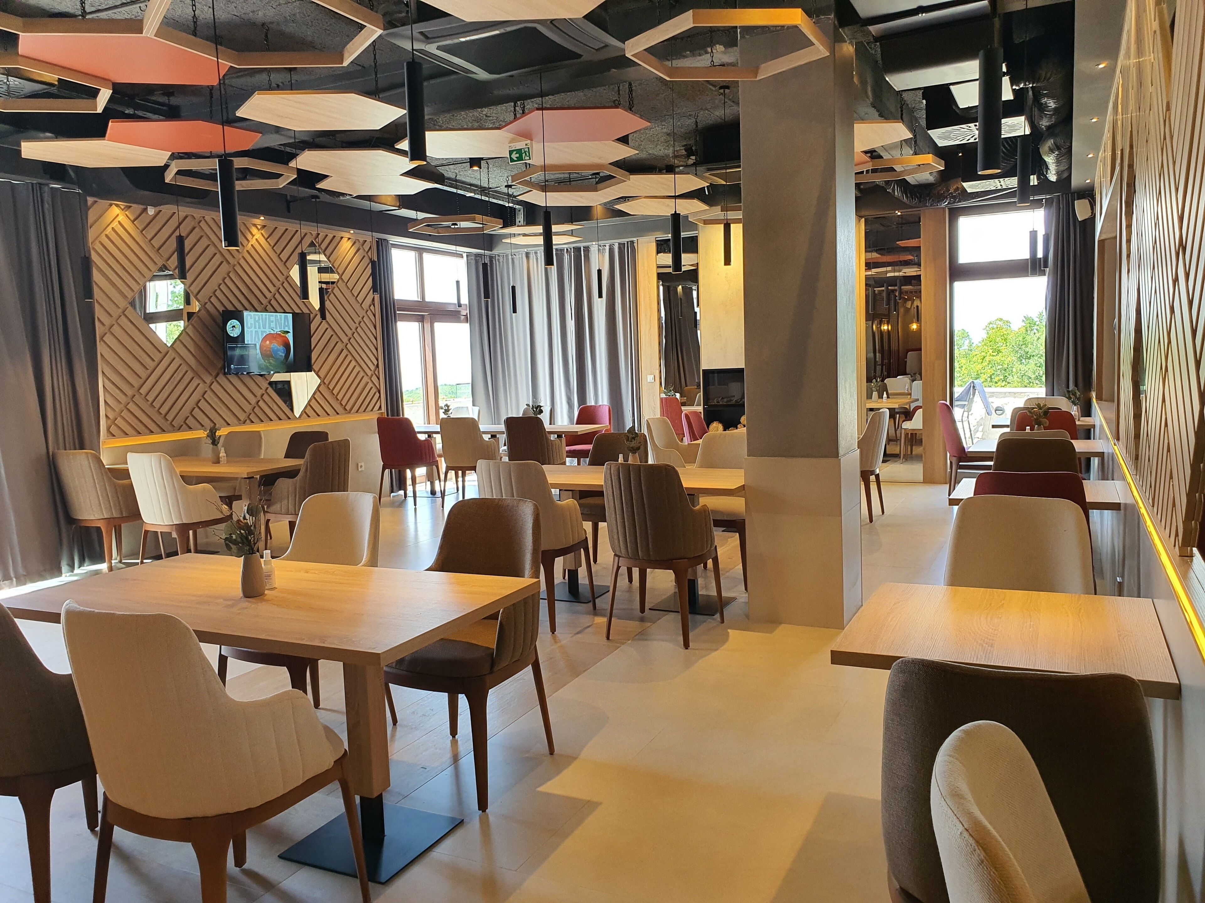A modern restaurant with wooden tables and chairs, a TV mounted on the wall, and large windows allowing natural light.