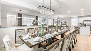 A modern dining area with a long table set for six, a chandelier, and a kitchen in the background.