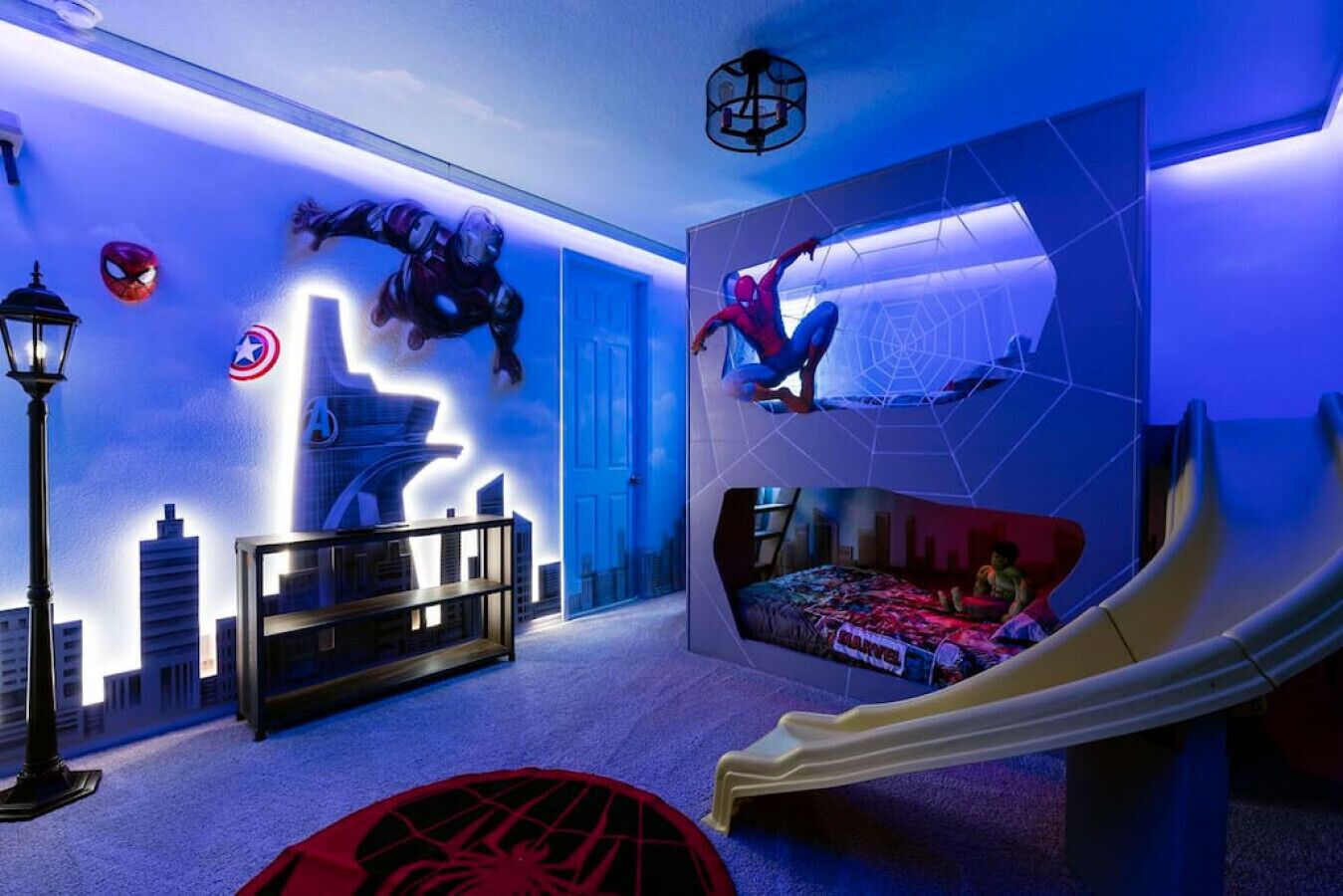 Une chambre avec un lit, un bureau, une chaise, une lampe et un mur décoré de motifs de super-héros.
