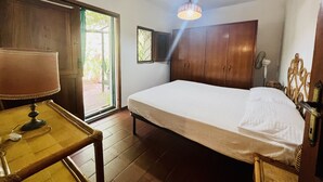 A bedroom with a bed, wooden wardrobe, a table with a lamp, and a window with curtains. - Trilocale in Centro con Giardino (San Teodoro)