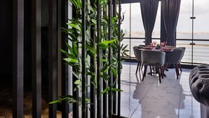 A modern living room with a large bamboo plant, a dining area with a table and chairs, and a view of the water through large windows. - ZAM-ZAM (Baku)