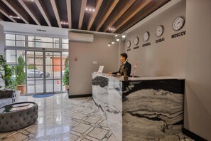 Zona de recepción de hotel con un mostrador de mármol, relojes que muestran la hora de diferentes ciudades y vistas a la calle.