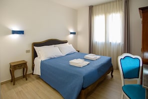 A neatly made bed with a blue bedspread, two white pillows, and folded towels on the side. There is a wooden nightstand with a lamp, a wooden chair with a blue cushion, and a window with curtains.