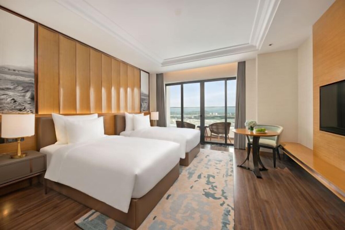 A hotel room with two beds, a large window, a TV, and a balcony with a view.